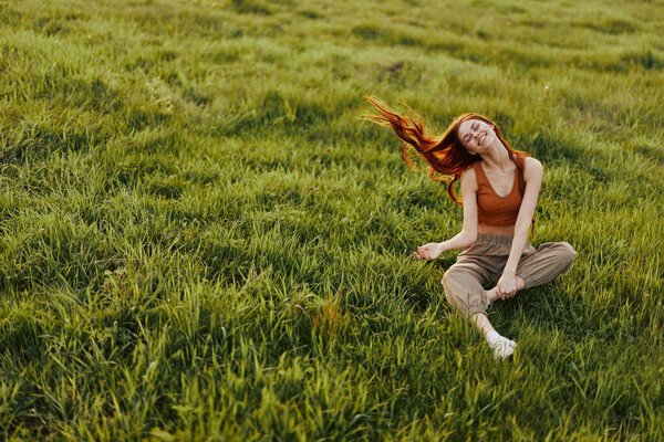 Young woman with red hair is happy and smiling sitting on the fresh grass in the park in the sun. High quality photo