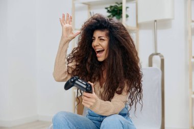Zevkten dört köşe olmuş, bronzlaşmış kıvırcık tenli Latin bayan iki elinde joystick oyun platformuyla 