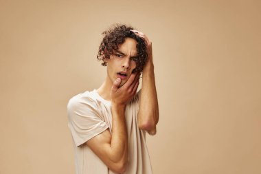 Temel tişörtlü harika bronzlaşmış kıvırcık adam, bej renkli pastel arka planda izole edilmiş pozlar veren yanaklara dokunuyor. Yeni Moda Koleksiyonu teklifi. İnsanlar ve Duygular konsepti. Reklam için boş yer