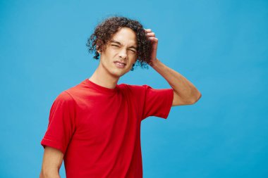 Düşünceli, düşünceli, mutsuz, bronzlaşmış, beyaz tenli, temel kırmızı tişörtlü adam mavi arka planda yalnız poz veriyor. Yaşam tarzı ve Duygular konsepti. Reklam için fotokopi alanı için iyi teklif.