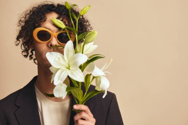 Güneş gözlüklü komik, yakışıklı, bronzlaşmış kıvırcık adam bej renkli pastel arka planda yalnız poz veren zambakları yüzüne yakın tutuyor. Yeni Moda Koleksiyonu teklifi. Retro stil konsepti. Reklam için boş yer