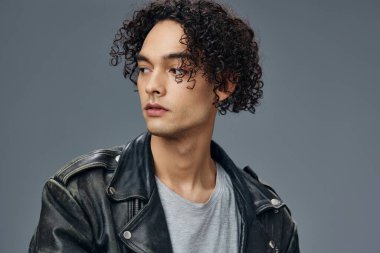 Dalgın, şık, bronzlaşmış, kıvırcık deri ceketli adam gri stüdyo arka planında yalnız poz veriyor. Güzel moda teklifi. Büyük Mevsimlik Satış Yeni Koleksiyon konsepti. İlan için alanı kopyala