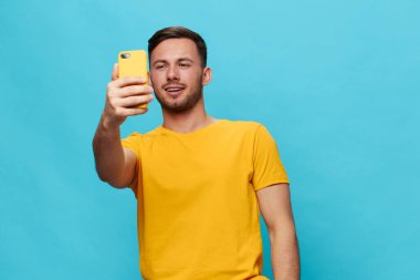 Sarı tişörtlü neşeli, bronzlaşmış yakışıklı adam mavi stüdyo arka planında izole edilmiş bir şekilde telefonuyla selfie görüntülü görüşme yapıyor. Uzayın afiş modelini kopyala. Çevrimiçi iletişim kavramı