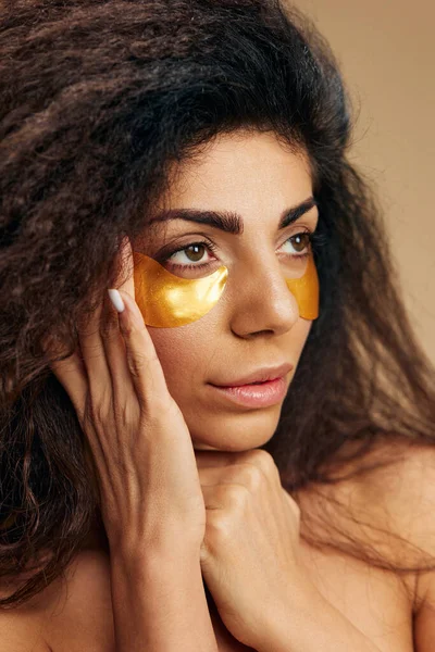 Pensive attractive Latin lady with gold hydrogel patches under eye touch facial mask look aside posing isolated over pastel beige background. Cosmetic product ad Natural beauty Skincare concept