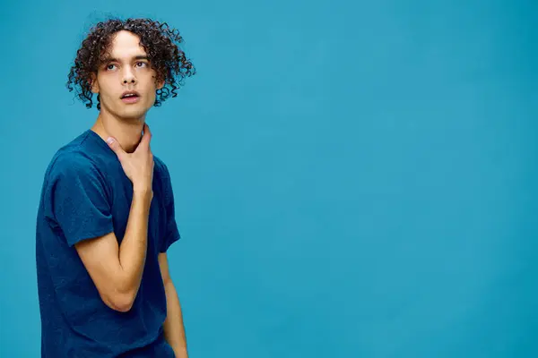 Confused pensive funny curly tanned Caucasian guy in basic t-shirt posing isolated on over blue background. Lifestyle and Emotions concept. Good offer with copy space for ad