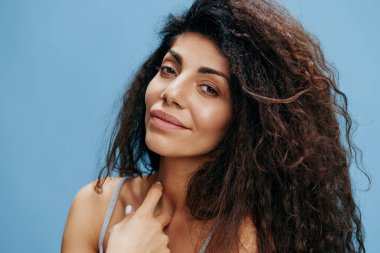 Vesikalık portre. Muhteşem kıvırcık Latin kadın izole edilmiş mavi gökyüzü arkaplanından zevk alıyor, kameraya bakın ve gülümseyin. Moda ve kozmetik markaları için iyi bir teklif. Boşluğu kopyala, reklam için boş yer