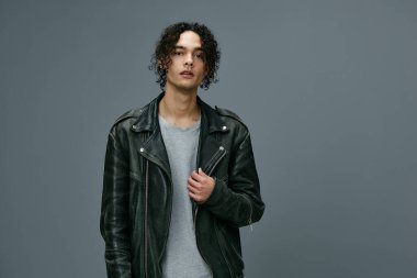 Kendine güvenen, bronz tenli, kıvırcık deri ceketli adam gri stüdyo arka planında izole edilmiş kameraya bakıyor. Güzel moda teklifi. Büyük Mevsimlik Satış Yeni Koleksiyon konsepti. İlan için alanı kopyala
