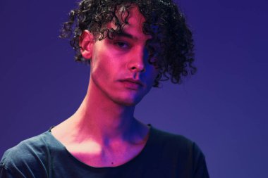 Siyah tişörtlü şık kıvırcık adam pembe mavi stüdyo arka planında yalnız poz veren kameraya bakıyor. Güzel moda teklifi. Sanal Gerçeklik Yeni Koleksiyon konsepti. İlan için alanı kopyala