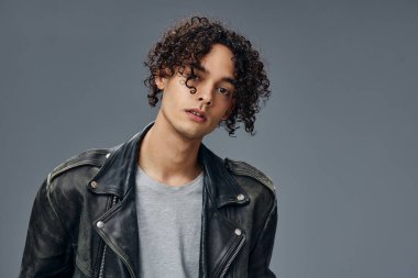 Ciddi bir şekilde bronzlaşmış kıvırcık deri ceketli adam portresi gri stüdyo arka planında izole edilmiş kameraya bakıyor. Güzel moda teklifi. Büyük Mevsimlik Satış Yeni Koleksiyon konsepti. İlan için alanı kopyala