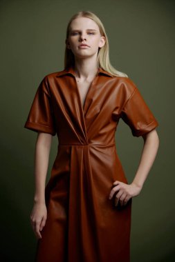 Markan için şık bir teklif. Modaya uygun sarışın kadın, el ele tutuşmuş deri yağmurluk giymiş, yeşil stüdyo arka planında izole edilmiş kameraya ciddi şekilde bakıyor.