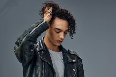 Yakışıklı, bronzlaşmış, kıvırcık deri ceketli adam saçlarına dokunuyor gri stüdyo arka planında izole bir şekilde poz veriyor. Güzel moda teklifi. Büyük Mevsimlik Satış Yeni Koleksiyon konsepti. İlan için alanı kopyala