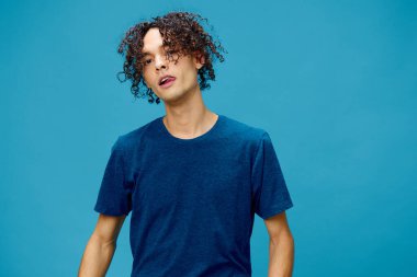 Narsistik, tapılası kıvırcık, bronzlaşmış, beyaz tişörtlü adam mavi arka planda yalnız poz veren kameraya bakıyor. Yaşam tarzı ve Duygular konsepti. Reklam için fotokopi alanı için iyi teklif.