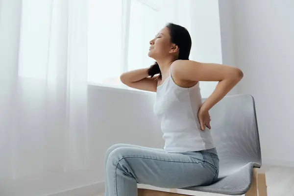 Back pain. Suffering from osteochondrosis after long study work pretty young Asian woman touching painful lower back at home interior living room. Injuries Poor health Illness concept. Cool offer