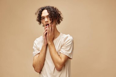 Temel tişörtlü harika bronzlaşmış kıvırcık adam, bej renkli pastel arka planda izole edilmiş pozlar veren yanaklara dokunuyor. Yeni Moda Koleksiyonu teklifi. İnsanlar ve Duygular konsepti. Reklam için boş yer
