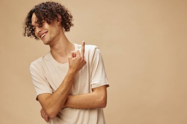 Temel tişörtlü, harika bronzlaşmış kıvırcık adam bej renkli pastel arka planda izole edilmiş orta parmak pozunu gösteriyor. Yeni Moda Koleksiyonu teklifi. İnsanlar ve Duygular konsepti. Reklam için boş yer