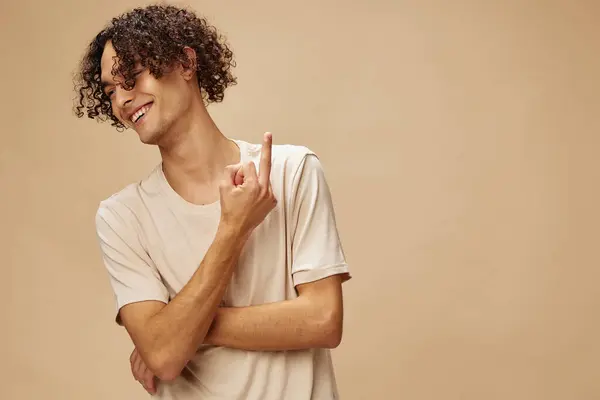 Defiant awesome tanned curly man in basic t-shirt show middle finger aside posing isolated on over beige pastel background. Fashion New Collection offer. People and Emotions concept. Free place for ad