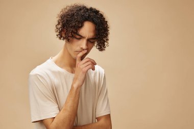 Basit tişörtlü komik, bronzlaşmış kıvırcık adam bej renkli pastel arka planda izole bir şekilde poz veremez. Yeni Moda Koleksiyonu teklifi. İnsanlar ve Duygular konsepti. Reklam için boş yer