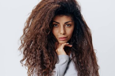 Beyaz arka plan stüdyosunda yalnız poz veren Afro kıvırcık saçlı, yüzünün yanındaki kameraya bakan kendine güvenen Latin kadının yakın portresi. Moda ve kozmetik markaları için iyi bir teklif.