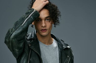 Seksi, şık, bronzlaşmış, kıvırcık deri ceketli adam gri stüdyo arka planında izole edilmiş kameraya bakıyor. Güzel moda teklifi. Büyük Mevsimlik Satış Yeni Koleksiyon konsepti. İlan için alanı kopyala