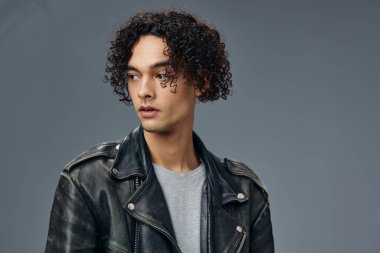 Dalgın, şık, bronzlaşmış, kıvırcık deri ceketli adam gri stüdyo arka planında yalnız poz veriyor. Güzel moda teklifi. Büyük Mevsimlik Satış Yeni Koleksiyon konsepti. İlan için alanı kopyala