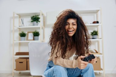 Keyifli, bronzlaşmış kıvırcık kıvırcık Latin bayanın iki elinde de joystickli oyun hamuru vardı. Kameraya gülümsüyor ve modern iç mekandaki sandalyeye oturuyordu. Uzayı kopyala Mockup Banner. Oyun platformunu kabul et