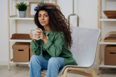 Hüsrana uğramış, bronzlaşmış, kıvırcık Latin bayan rahat gömlekli çay kahvesiyle sorunu olan ev yapımı modern arka plandaki sandalyeye oturup kameraya bakın. Uzay Sancağını kopyala. Kavram psikolojisi