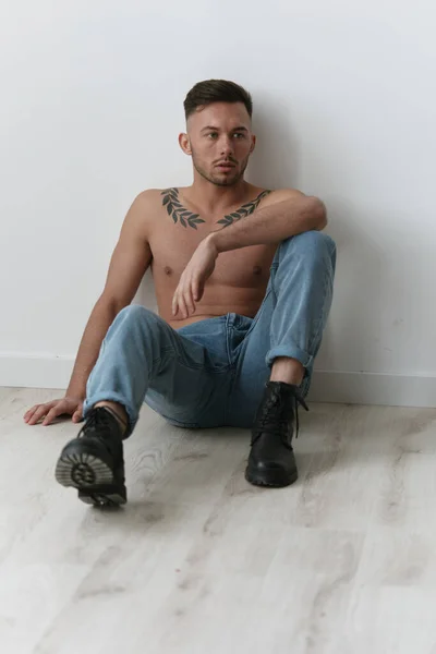 Modelling snapshots. Serious attractive naked handsome man in jeans sitting on floor posing in white studio background. Fashion Men Shoot concept. Copy space. Cool offer for ad