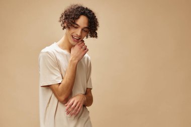 Temel tişörtlü neşeli, harika, bronzlaşmış kıvırcık adam bej renkli pastel arka planda yalnız poz veriyor. Yeni Moda Koleksiyonu teklifi. İnsanlar ve Duygular konsepti. Reklam için boş yer