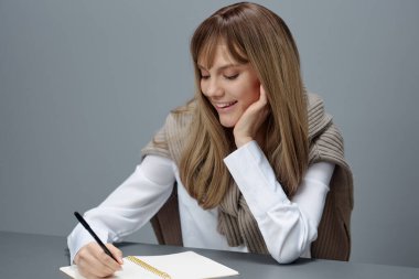 Happy young blonde student journalist lady in warm sweater writing love letter article essay sitting at workplace in gray modern home office. Education And Career Profession Concept. Copy space clipart