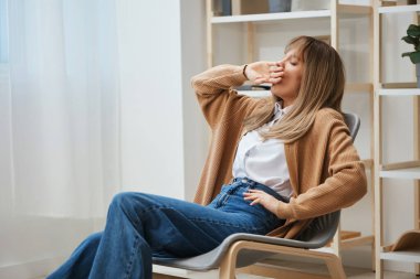 Uykulu, yorgun, sarışın, sıcak süveter giyen, esneyen ve evinde elini koltuğuna koyup ağzını kapatan bir bayan. İşten ara vermek, ara vermek, boş zaman kavramını yansıtmak. Boşluğu kopyala