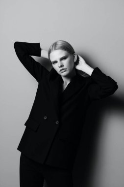 Klasik takım elbiseli çekici sarışın bir kız elleriyle saçlarını tutuyor ve stüdyoda siyah beyaz tarzda profesyonel bir model gibi poz veriyor. Moda fotoğrafçılığı. Kıyafet mağazaları için konsept. Yüksek kalite fotoğraf
