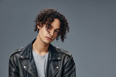 Kendine güvenen, bronz tenli, kıvırcık deri ceketli adam gri stüdyo arka planında izole edilmiş kameraya bakıyor. Güzel moda teklifi. Büyük Mevsimlik Satış Yeni Koleksiyon konsepti. İlan için alanı kopyala