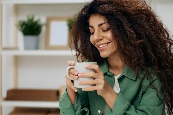 Cute smiling enjoyed pretty tanned curly Latin lady look with love aside drink tea coffee in casual shirt sit on chair in home modern interior background. Copy space Banner. Nostalgia concept