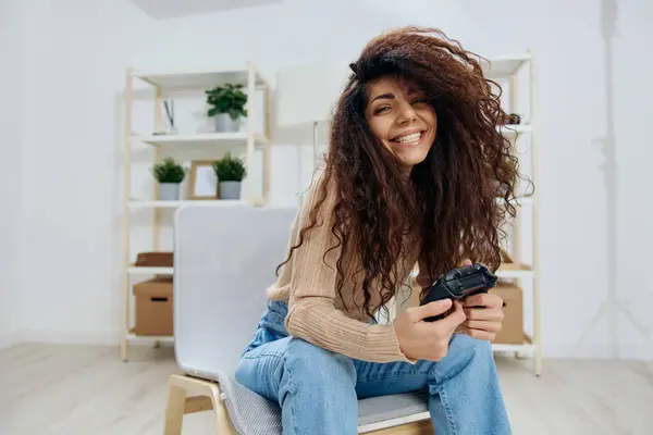 Smiling pretty enjoyed tanned curly Latin winner lady hold with joystick gamepad in both hands sit on chair in home modern interior look at camera. Copy space Mockup Banner. Concept game platform