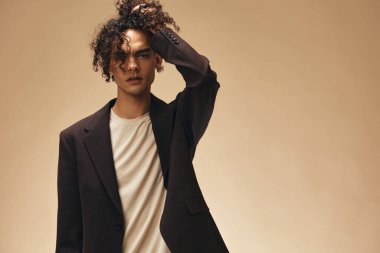 Klasik kahverengi ceketli, bronzlaşmış muhteşem kıvırcık adam bej renkli pastel arka planda izole edilmiş buklelere dokunuyor. Yeni Moda Koleksiyonu teklifi. Retro stil konsepti. Reklam için boş yer