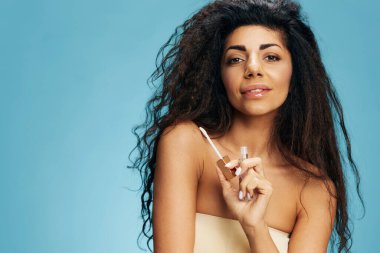 Neşeli, seksi, kıvırcık kıvırcık, kıvırcık, dudak parlatıcısı olan Latin kadın pastel mavisi arka planda izole edilmiş poz veren kameraya bak. Kozmetik dudak ürünü. Doğal güzellik konsepti. Stüdyo portresi.