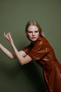 Deri kahverengi elbiseli zarif sarışın kadın zeytin yeşili stüdyo arka planında yalnız poz veriyor. Yeni koleksiyon afişi için havalı bir moda teklifi