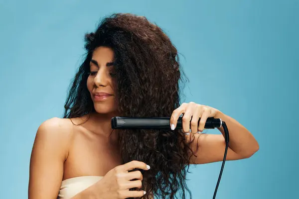 Güzel Latin Kıvırcık Kadın Saç Düzleştirici Kullanıyor Aşağıya Bakıyor Mavi — Stok fotoğraf