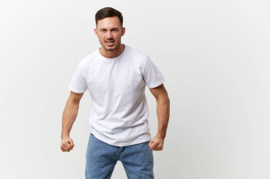 Çığlık atan, kızgın, bronzlaşmış, basit tişörtlü yakışıklı adam beyaz stüdyo arka planında izole edilmiş yumruklarını sıkıyor. Uzayın afiş modelini kopyala. İnsanların duyguları Yaşam tarzı konsepti