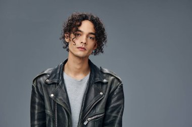 Oldukça seksi, kendine güveni olan, bronzlaşmış, kıvırcık deri ceketli, gri stüdyo arka planında yalnız poz veren bir adam. Güzel moda teklifi. Büyük Mevsimlik Satış Yeni Koleksiyon konsepti. İlan için alanı kopyala