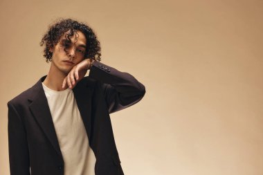 Klasik kahverengi ceketli, bronzlaşmış muhteşem kıvırcık adam bej renkli pastel arka planda izole edilmiş buklelere dokunuyor. Yeni Moda Koleksiyonu teklifi. Retro stil konsepti. Reklam için boş yer