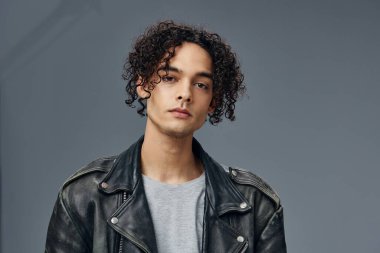 Ciddi bir şekilde bronzlaşmış kıvırcık deri ceketli adam portresi gri stüdyo arka planında izole edilmiş kameraya bakıyor. Güzel moda teklifi. Büyük Mevsimlik Satış Yeni Koleksiyon konsepti. İlan için alanı kopyala