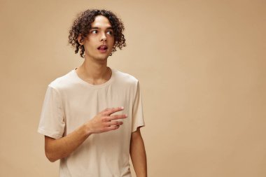 Esnek tişörtlü bronzlaşmış harika kıvırcık adam bej renkli pastel arka planda yalnız poz veriyor. Yeni Moda Koleksiyonu teklifi. İnsanlar ve Duygular konsepti. Reklam için boş yer