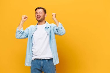 Mavi tişörtlü mutlu, bronz tenli yakışıklı adam yumruklarını kaldırıp turuncu sarı stüdyo arka planında poz verme konusunda çok şanslı. Uzayın afiş modelini kopyala. İnsanların duyguları Yaşam tarzı konsepti