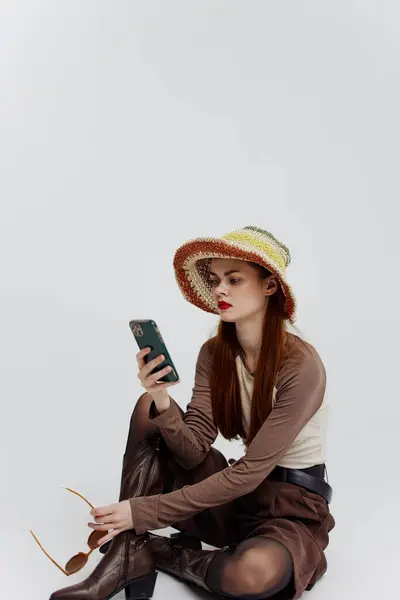 一位戴帽子的专心致志的女士坐在一间白色工作室的地板上 一边打电话一边发短信 示范目录摄影 高质量的照片 — 图库照片
