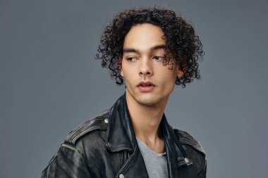 Dalgın, şık, bronzlaşmış, kıvırcık deri ceketli adam gri stüdyo arka planında yalnız poz veriyor. Güzel moda teklifi. Büyük Mevsimlik Satış Yeni Koleksiyon konsepti. İlan için alanı kopyala