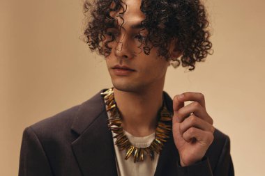 Klasik kahverengi ceketli harika bronzlaşmış kıvırcık adam bej renkli pastel arka planda izole edilmiş etnik kolyeye dokunuyor. Yeni Moda Koleksiyonu teklifi. Retro stil konsepti. Reklam için boş yer