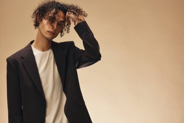 Klasik kahverengi ceketli, bronzlaşmış muhteşem kıvırcık adam bej renkli pastel arka planda izole edilmiş buklelere dokunuyor. Yeni Moda Koleksiyonu teklifi. Retro stil konsepti. Reklam için boş yer