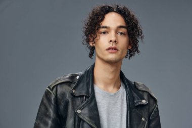 Oldukça seksi, kendine güveni olan, bronzlaşmış, kıvırcık deri ceketli, gri stüdyo arka planında yalnız poz veren bir adam. Güzel moda teklifi. Büyük Mevsimlik Satış Yeni Koleksiyon konsepti. İlan için alanı kopyala