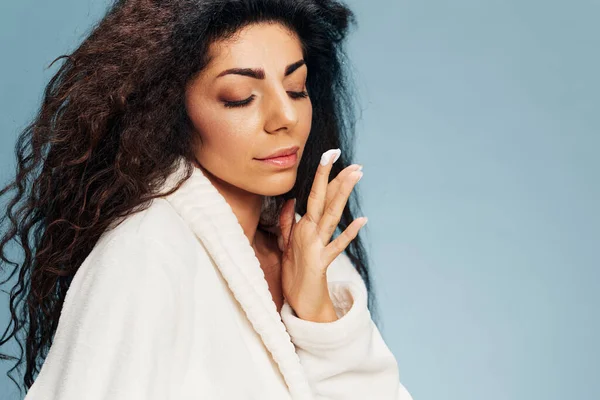 Pensive curly Latin lady in bathrobe looking at drop of cream on finger near perfect clean face, closing eyes, posing isolated on pastel blue background. Cosmetic product ad concept. Copy space